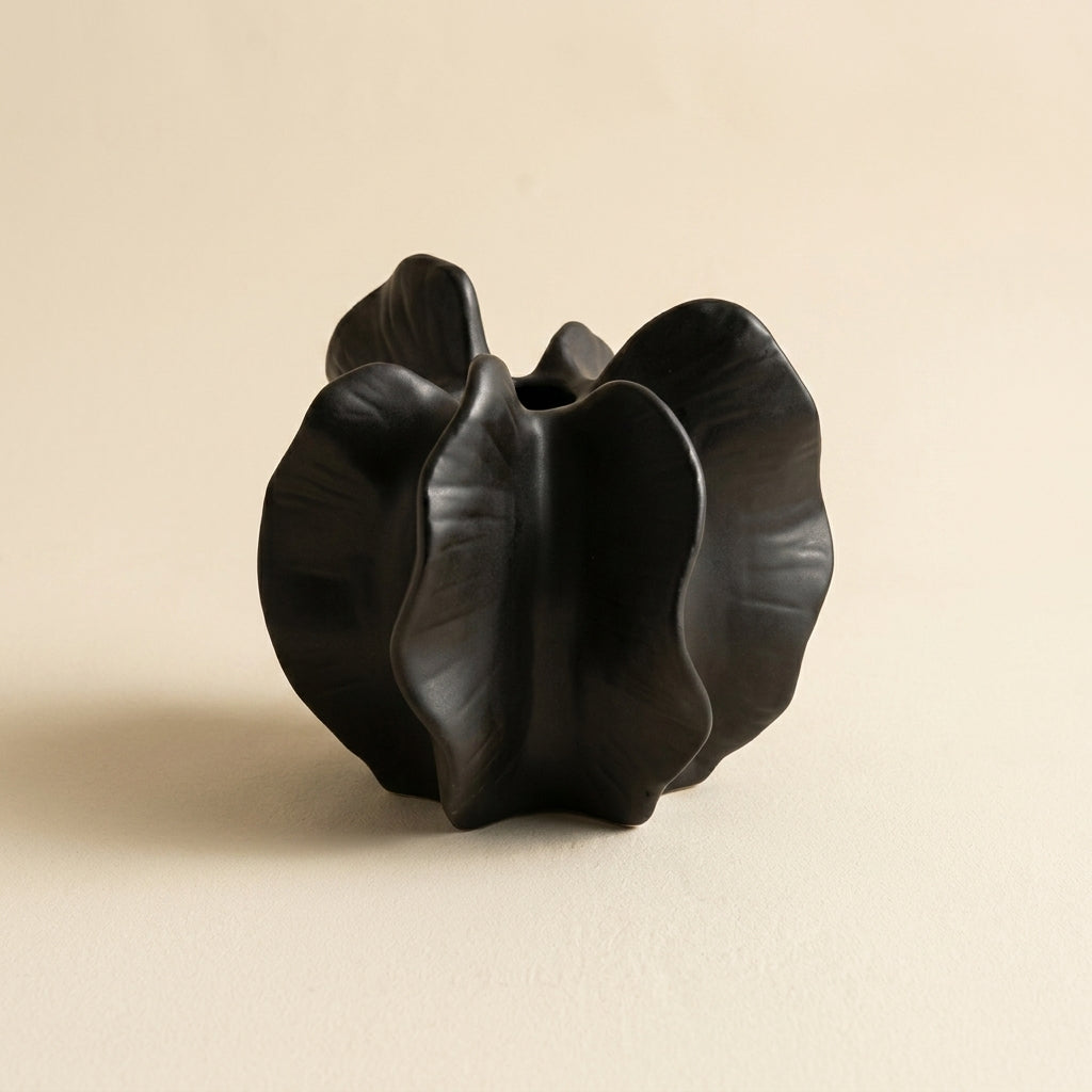 Matte black speckled ceramic wabi-sabi bloom vase with ruffled petal lobes and central cleft, on a warm-white studio backdrop