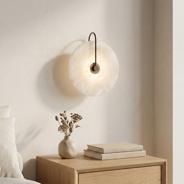 Alabaster-look moon disc sconce glowing warmly above a wood nightstand beside a bed