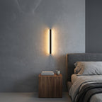 Short black linear LED bar wall light mounted vertically beside a bed and nightstand, warm glow