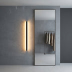Short black linear LED bar wall light beside a full-length mirror in a dressing area, warm glow