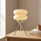 Two-tier cream glass macaron table lamp on a polished chrome tripod base, glowing warmly on a wooden side table.