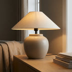 Wabi-sabi ceramic-look table lamp glowing warm beside books in a calm neutral bedroom setting.