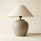 Matte oatmeal ceramic-look table lamp with cream coolie shade casting warm ambient light in a reading nook.