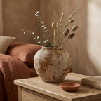 Round rustic wabi-sabi ceramic pot vase with dried stems on a bedroom nightstand